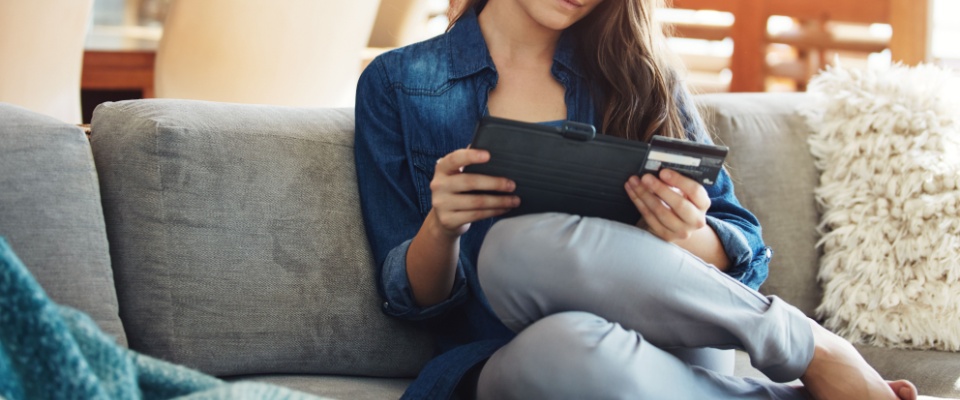 A woman sitting on a sofa at home, smiling as she reads information about her credit score on a tablet. She holds a card in her hand, symbolizing digital convenience and personal financial management in a comfortable domestic setting.