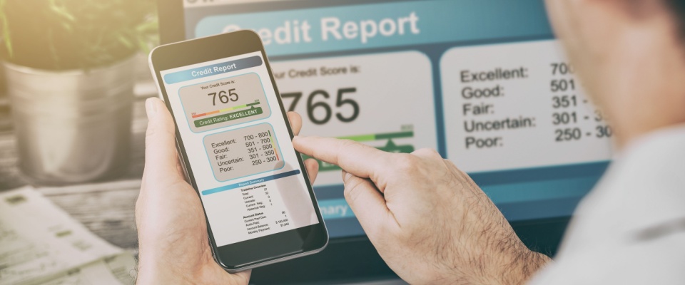 A person uses their smartphone to check on their credit score report. Their laptop in the background is also displaying their credit score information.