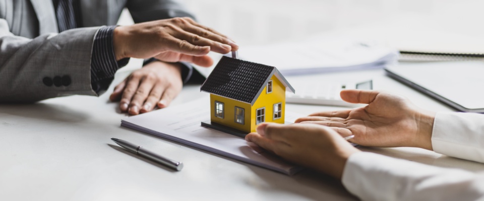 Two people negotiating regarding a housing contract, with a physical house model sitting on top of the paperwork for visualization purposes.