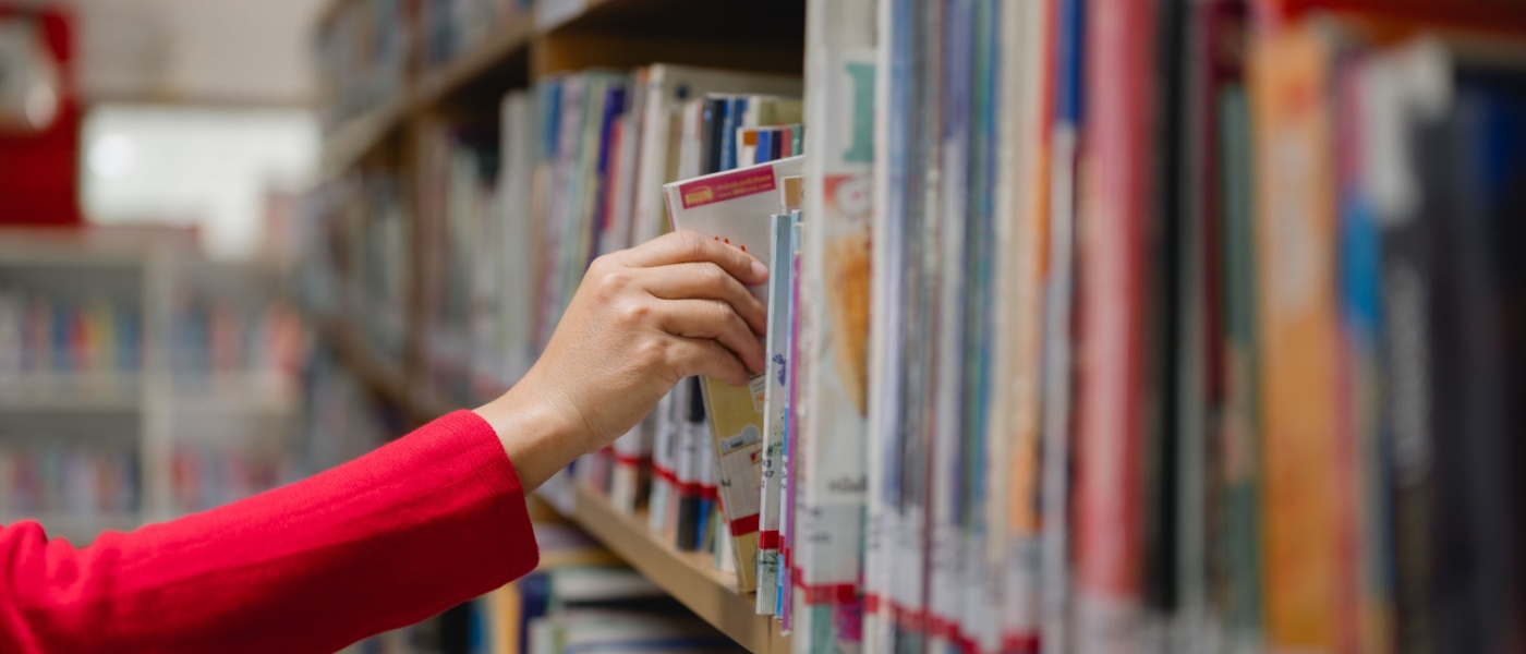 A woman in a library reaching for a book on a high shelf, surrounded by rows of books.