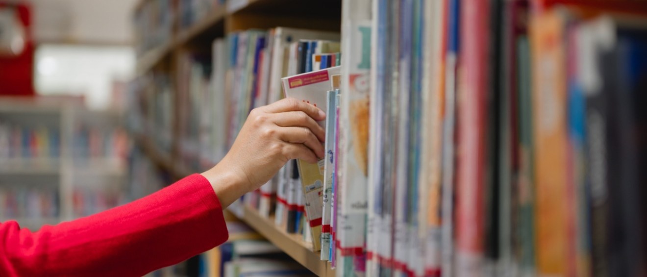 A woman in a library reaching for a book on a high shelf, surrounded by rows of books.