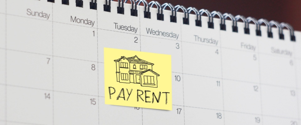 A close-up of a calendar, which has a sticky note on it, containing a drawing of a house and the words “Pay rent” written in all caps.