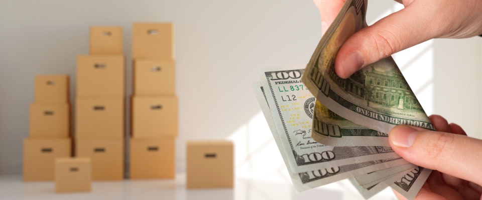 ands counting dollar bills with a background of stacked moving boxes, representing the hidden costs of moving in or out of an apartment.