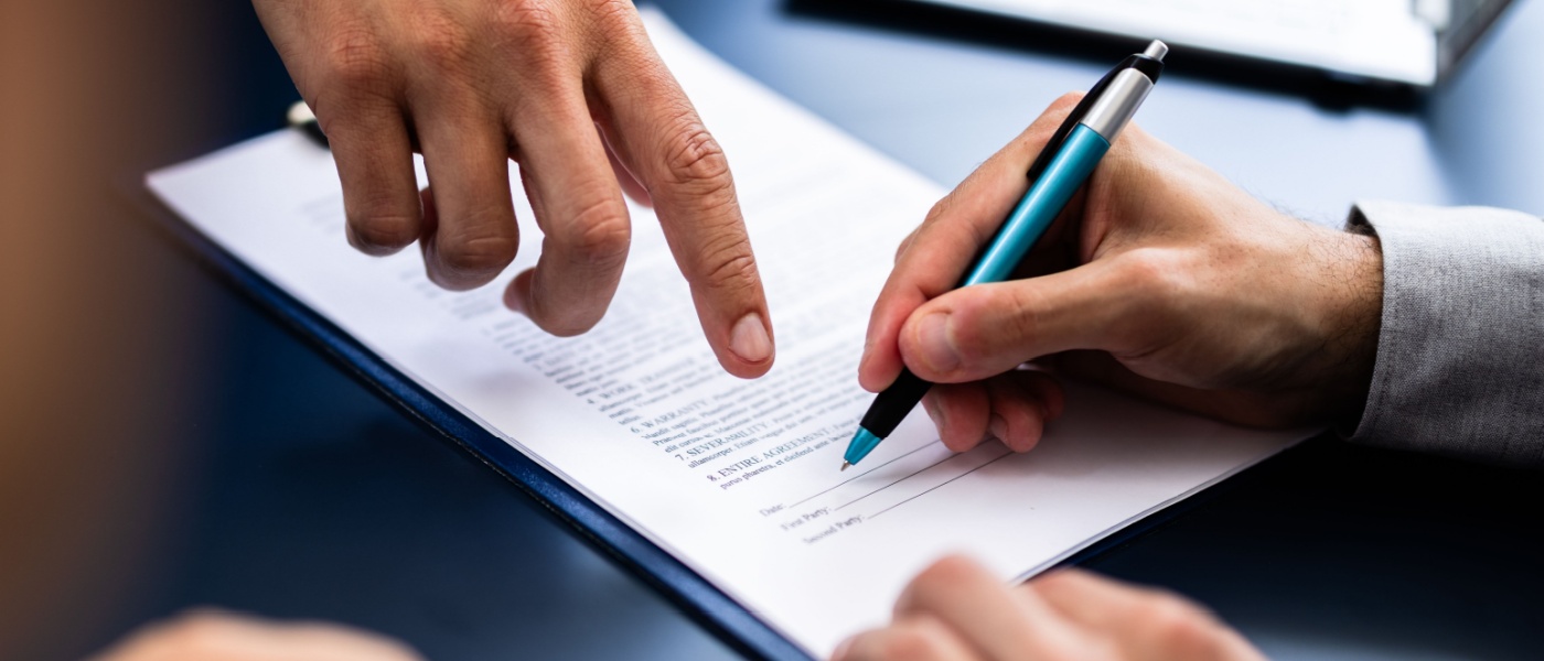 A close-up of a person, holding a pen in their right hand, about to sign a contract, while another person uses their right hand to point to the place where the paperwork should be signed.
