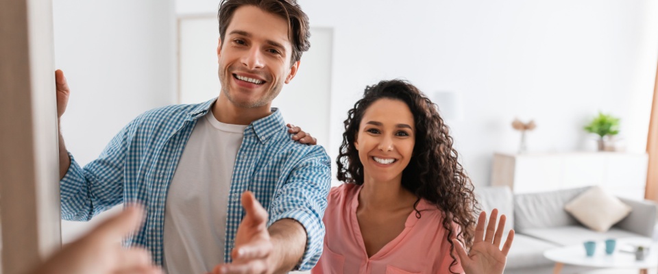 A cheerful couple standing in the doorway of their modern flat, smiling and inviting guests inside. 