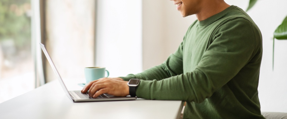 A side view of a stylish and cheerful young man working in a cozy cafe. He is drinking coffee while typing on a laptop keyboard, engaged in tasks like sending emails or coding. 