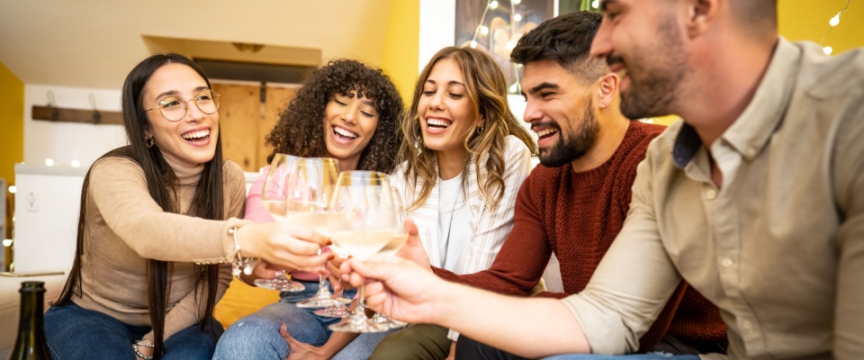 A group of multiracial young friends sitting on a sofa at home, toasting with champagne glasses in hand. 