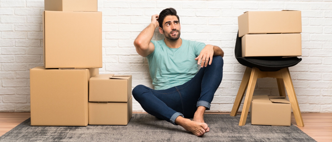 Young man moving in new home among boxes having doubts, with a confused face expression.