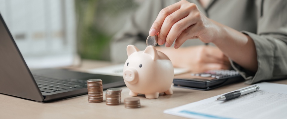 A woman placing coins into a piggy bank, symbolizing financial concepts such as loans, debt, savings, and wealth. A calculator and coins are nearby, emphasizing themes of tax planning, fund growth, profit, and retirement savings.