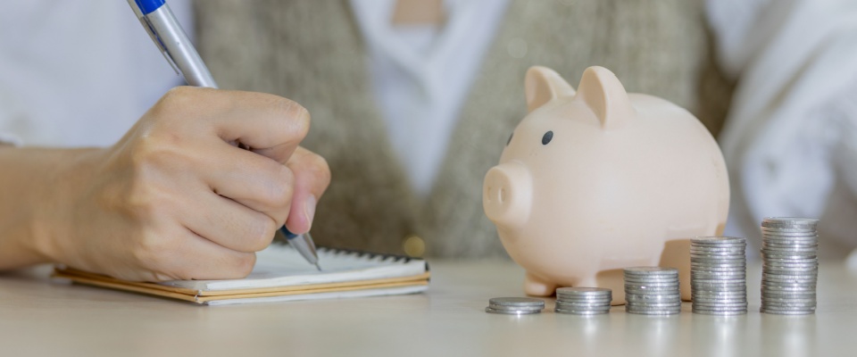 A woman’s hand placing a coin into a piggy bank, symbolizing saving money and financial planning. Next to the piggy bank, stacks of coins are neatly arranged, emphasizing concepts of wealth, budgeting, and economic growth.