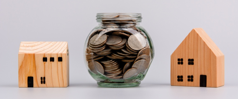 Jar filled with coins placed between two wooden house models, symbolizing security deposits as a hidden cost of renting.