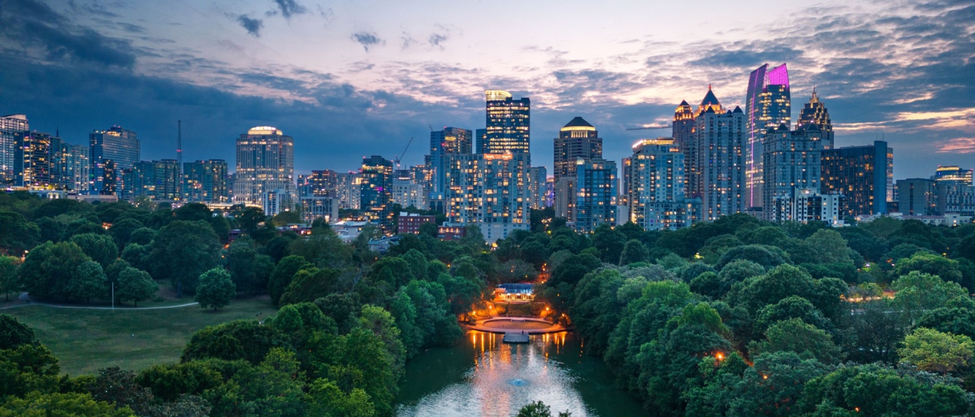 A view of the skyline in Atlanta, pictured at dusk, with its skyscrapers lit up against the darkening sky, and Piedmont Park visible in the front.