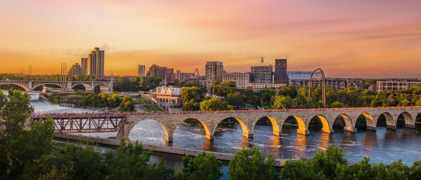 Sunset view of downtown Minneapolis, MN, featuring the Stone Arch Bridge, Mississippi River, and the city skyline.