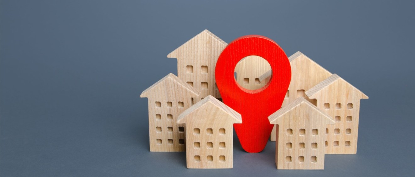 Wooden miniature houses with a large red location pin marker against a gray background simbolizing the concept of address change for renters moving to a new place.