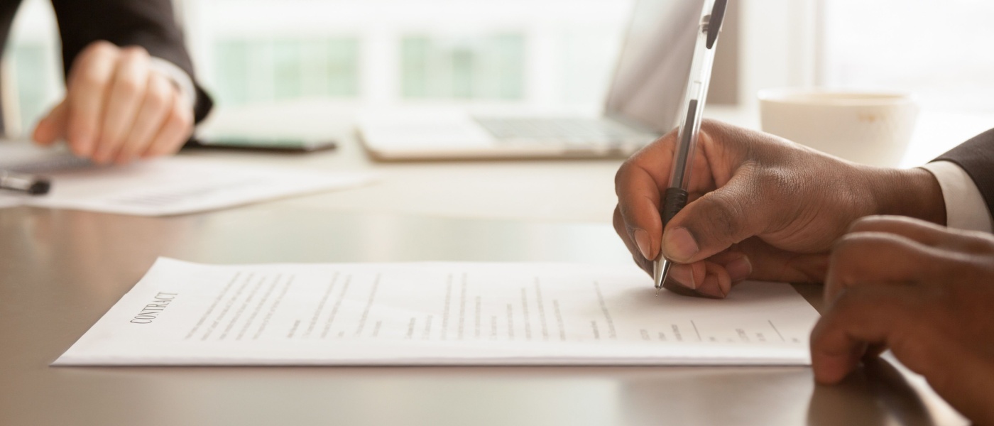 A close-up of a person, holding a ballpoint pen in their right hand, about to sign a contract, with another person’s hand being visible in the background.