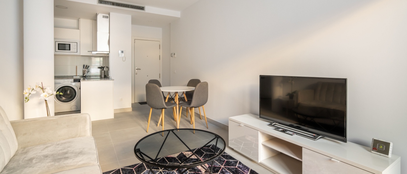 A photo of a studio apartment’s main living area, including a kitchenette, a dining table with four chairs around it, and a couch facing the TV in the living area.