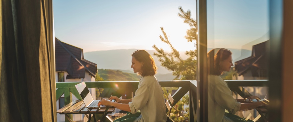 Young woman sitting on balcony with view of the mountains and the sunset, working and using a laptop in a garden-style apartment building.