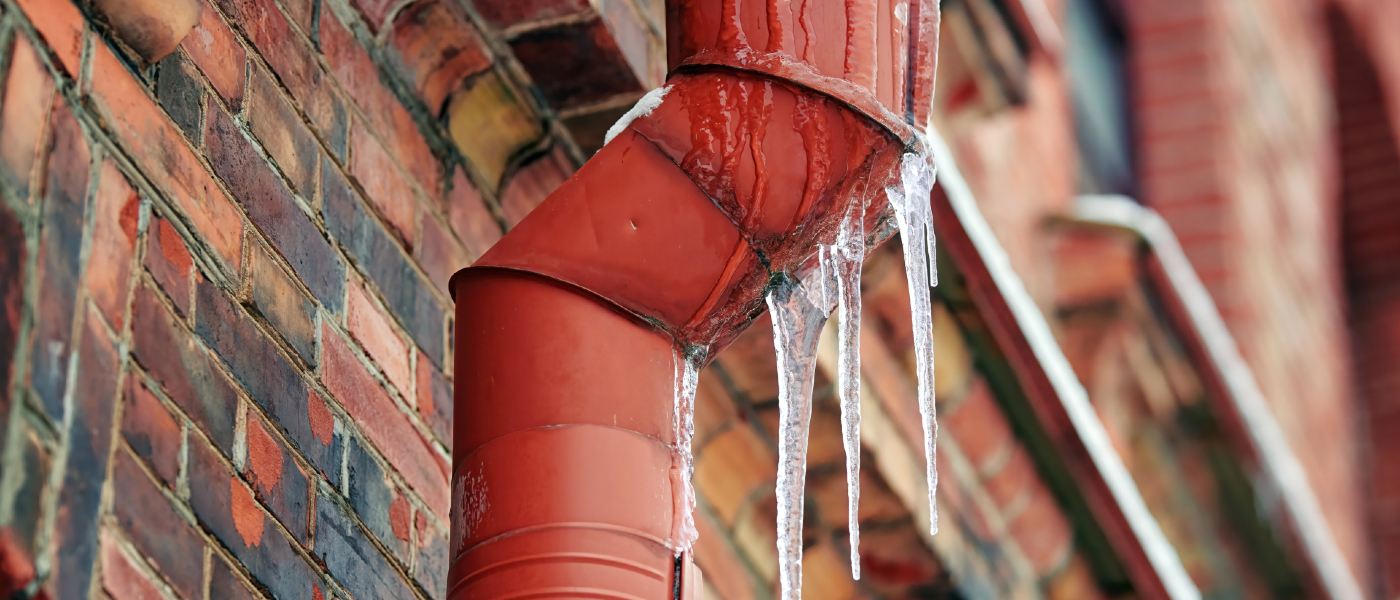 Red frozen pipe with icicles hanging from a bent section, attached to a red brick building during winter.