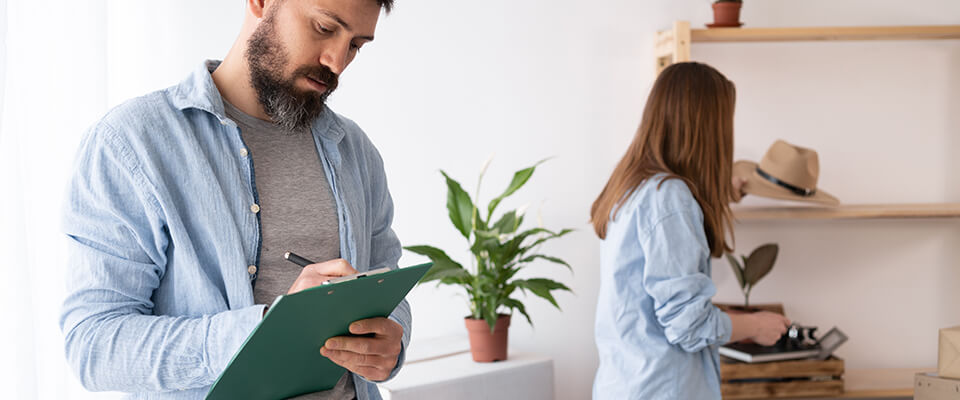 A man wearing a light blue shirt, holding a pen and writing in a green clipboard, and a red-headed woman wearing a light blue shirt, looking at a beige hat on a shelf.