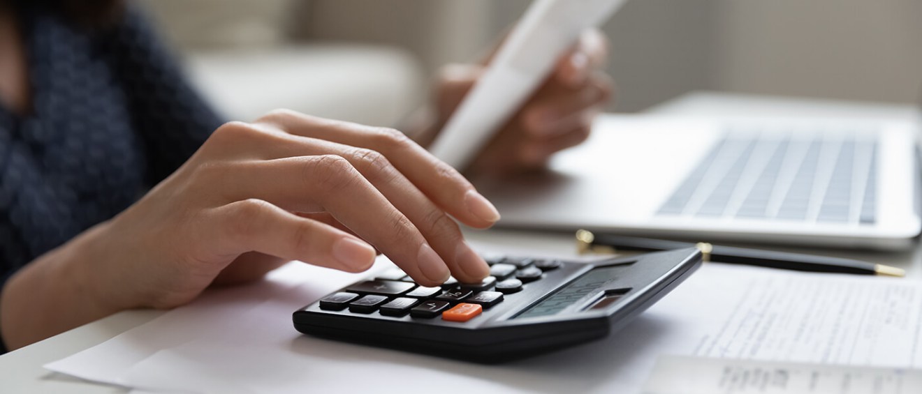 A woman using a calculator while reviewing financial documents at a desk, with a laptop and a pen in the background.