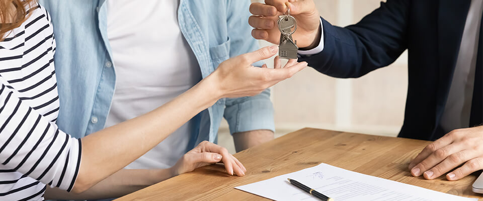A couple receiving the keys to an apartment from a real estate agent, sitting at a wooden table with a document and pen on it.