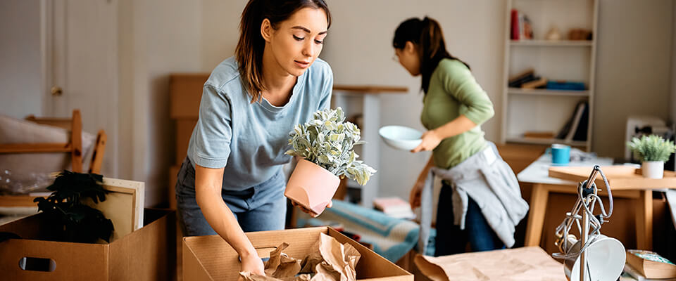 Two women unpacking boxes with plants and dishes and organizing belongings after moving into a new apartment.