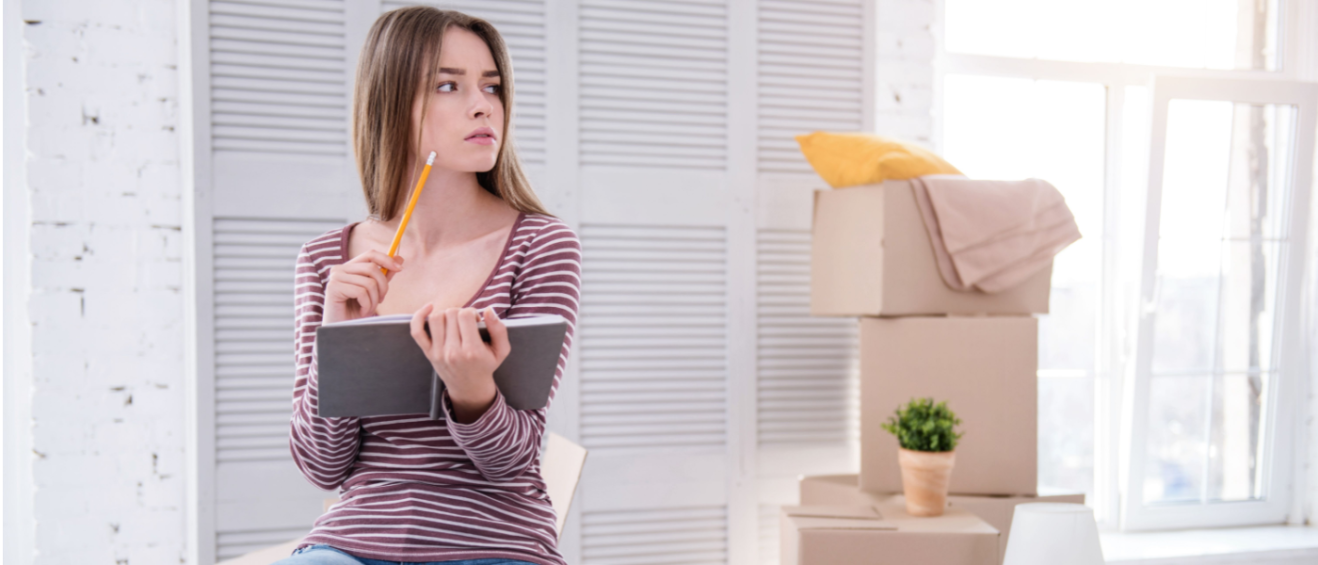 A woman in a striped shirt and jeans sitting in a bright apartment, holding a notebook and pencil while surrounded by stacked moving boxes.