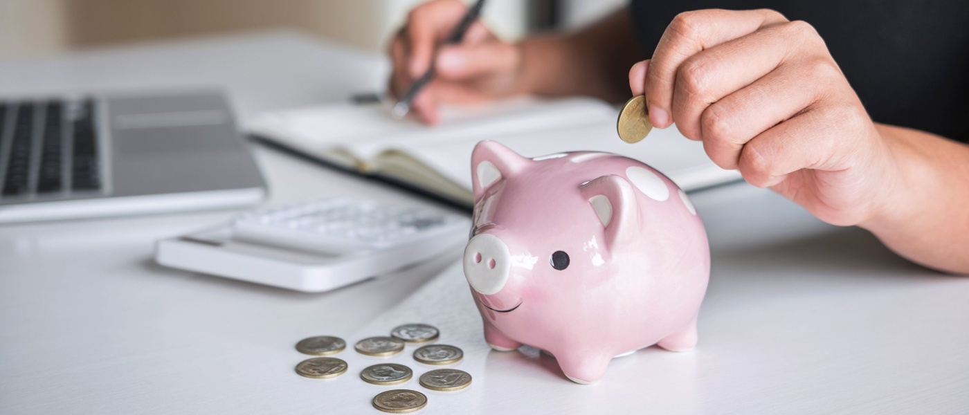 A person writes something in a notebook, while inserting coins into a pink porcelain piggy bank.