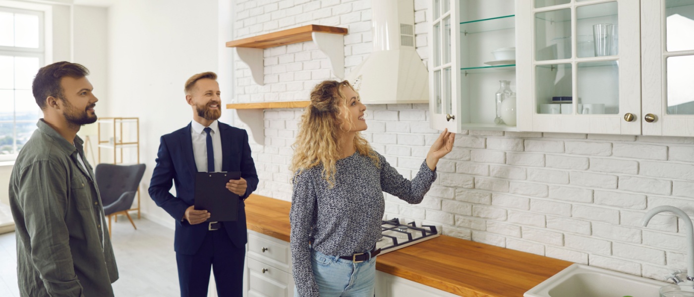 Future tenants or first time buyers meeting real estate agent and doin an apartment tour. Boyfriend and girlfriend buying new home. Husband and wife looking at modern white wooden cupboards in kitchen.