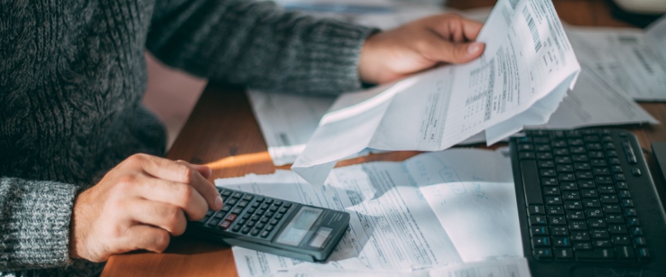 Close-up of a man's hands holding a utility bill, surrounded by multiple receipts and a calculator on the table, calculating gas, electricity, and heating costs amid rising service tariffs.