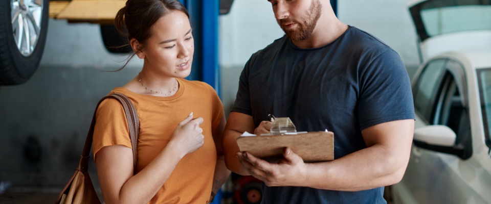Woman mechanic holding a clipboard and discussing a car maintenance invoice with a customer in an auto repair shop.