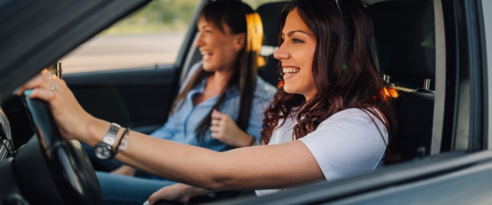 Smiling, fashionable female friends driving on a city road. Side view of a trendy woman gripping the steering wheel while her best friend sits beside her, laughing and enjoying the ride.