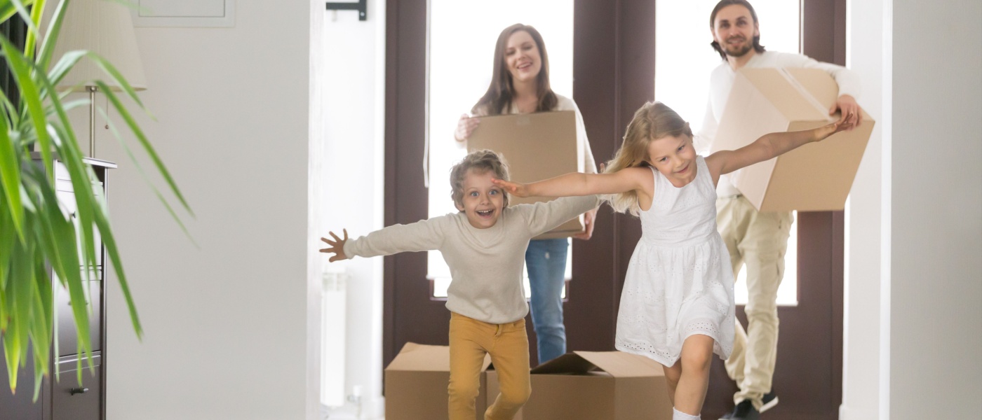 Happy married couple with young children arriving at their new modern home. Husband and wife carrying cardboard boxes while watching their excited daughter and son run inside.