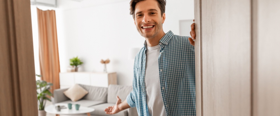 Cheerful man standing in the doorway of his modern apartment, smiling and inviting a visitor inside, gesturing towards the living room with his hand.