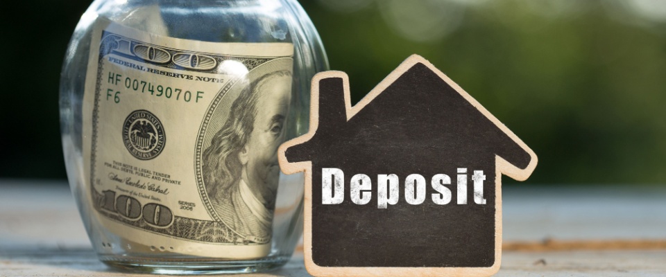 A $100 bill stuck inside of a jar, with the word “deposit” written on a house-shaped card next to the jar.