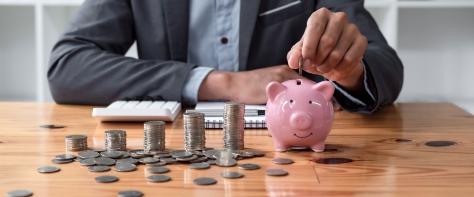 A person inserting a coin inside of a pink porcelain piggy bank, with stacks of coins next to it, and a calculator, notepad and pen behind it.