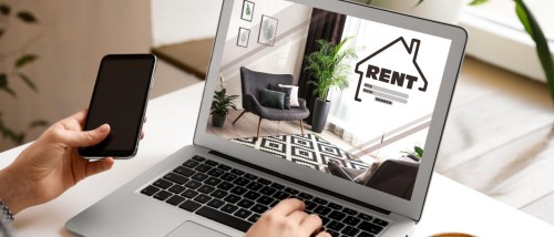 A person, using a laptop to search for an apartment for rent, while holding their smartphone in their left hand.