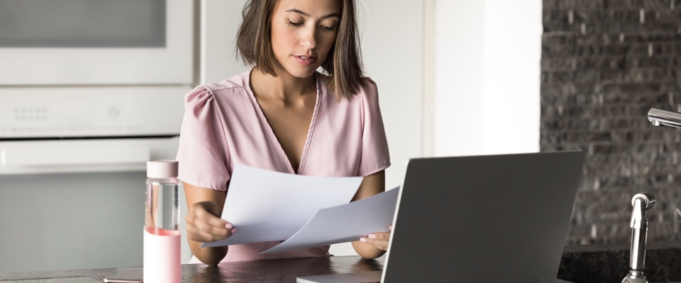 A 25-year-old woman sits at the kitchen counter with a laptop, focused on reviewing important documents, managing her budget, and working as a freelancer in the context of student loans.