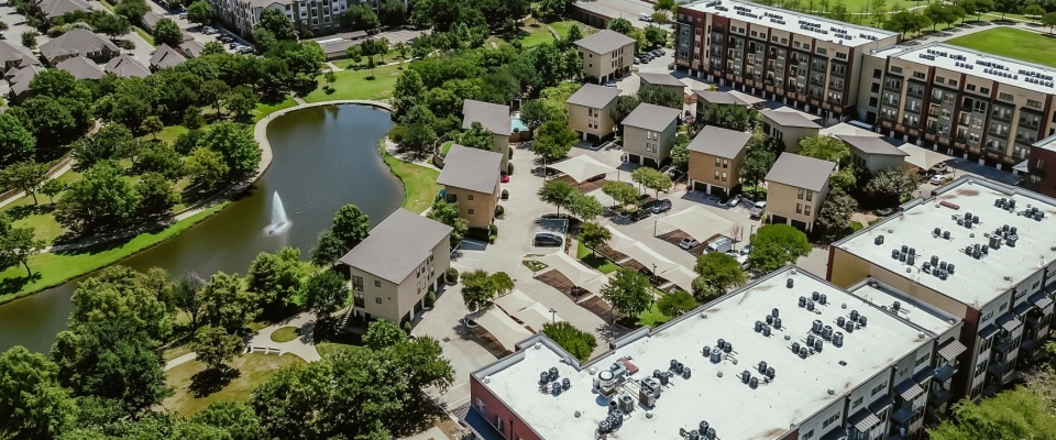 Aerial panoramic view of an urban park and apartment building with a shingle roof, flat roof with industrial rooftop AC units and furnace units in a steel cabinet, lush green park with pond and trails, Keller, Texas, USA.