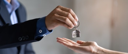 A landlord fulfilling their duties by handing over a house-shaped keychain to a tenant, who reaches out to receive it.