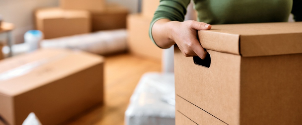 A renter moving in or out, carrying a cardboard box, with a stack of boxes in the background as part of the landlord duties of managing turnovers.
