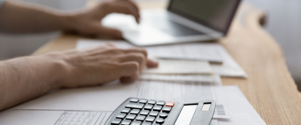 A person in the process of proving income when self-employed uses a calculator and a laptop to keep track of papers.