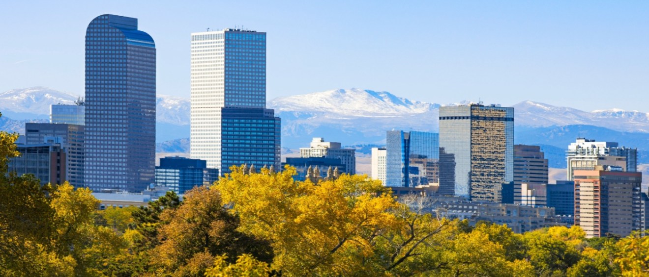 The Denver skyline, with autumn foliage and snow-capped mountains in the background, showcasing the city’s beauty and appeal for renters researching the cost of living in Denver.
