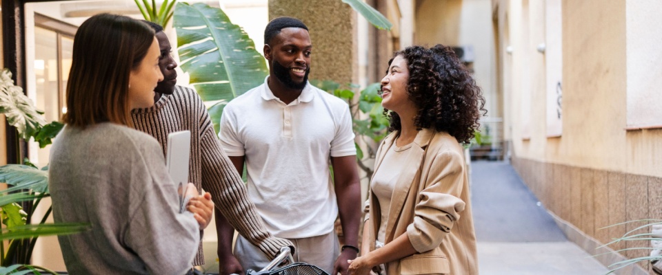 People talking and smiling in the hallway of a modern apartment building with plants and a bicycle, highlighting opportunities to ask questions for neighbors.