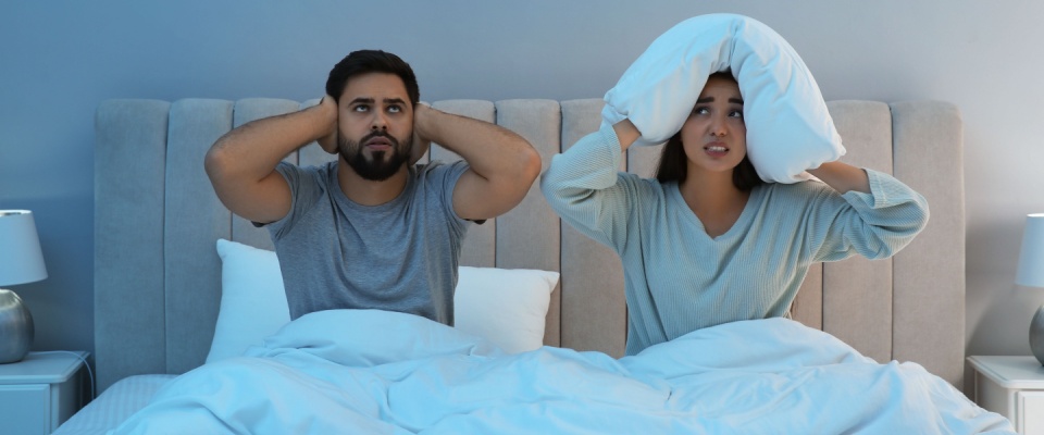 Frustrated couple in bed covering their ears, illustrating the importance of asking about noise insulation before renting an apartment in Denver.
