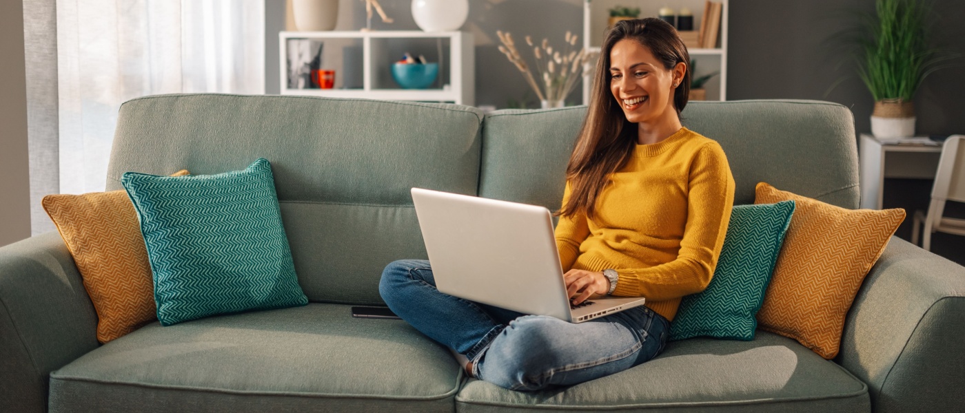 Cheerful young woman sitting on the sofa in her modern apartment, smiling while browsing online—possibly searching for a great deal on a rental or exploring new opportunities from the comfort of home.
