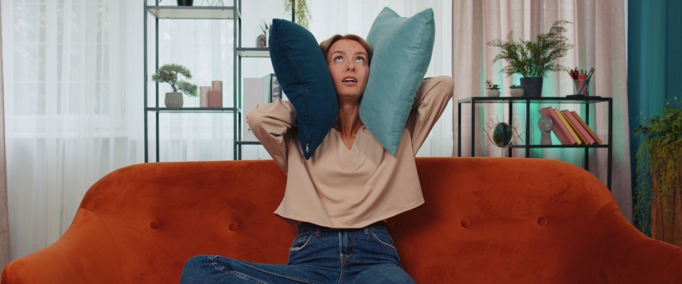 Irritated woman sitting on a couch, covering her ears with pillows to block out noise from repair work next door—illustrating the challenges of living in a flat with thin walls and poor sound insulation, often a concern in apartments in Chicago.