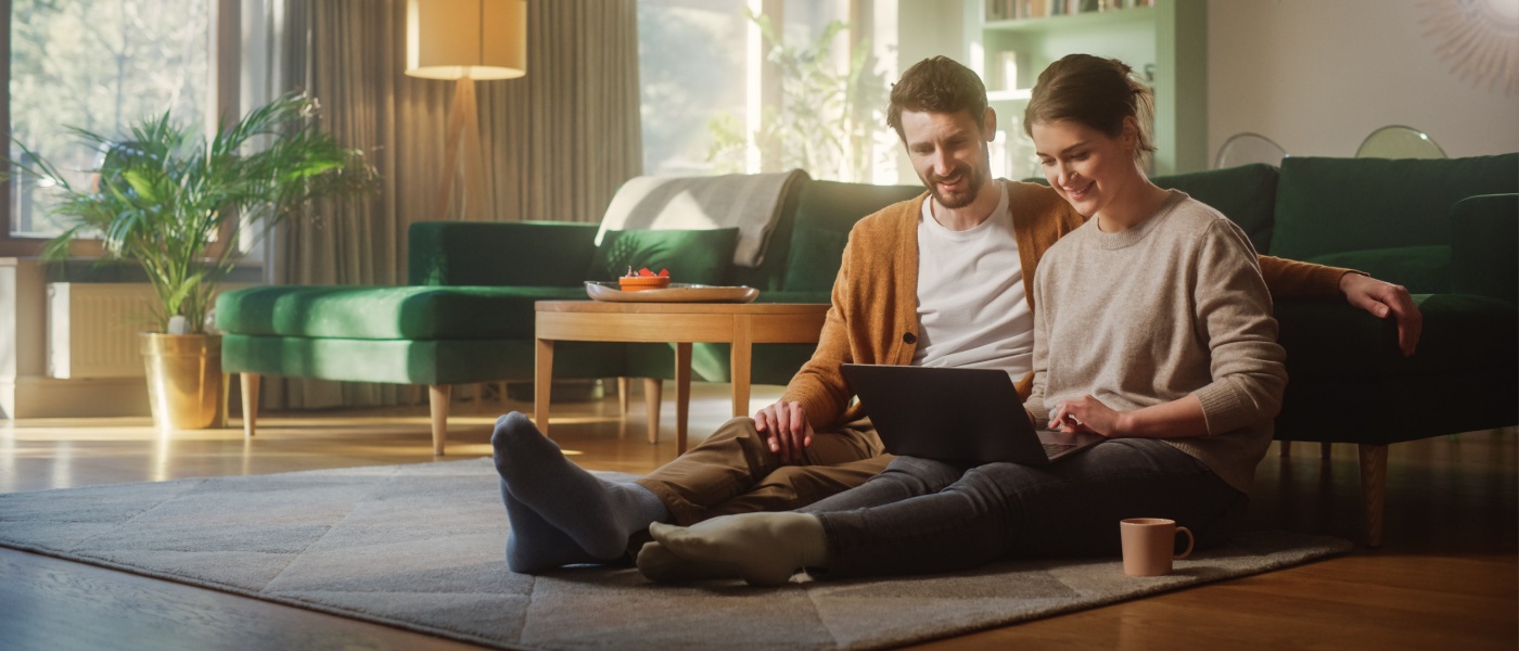 Couple sitting on the living room floor of their apartment using a laptop together—engaged in their apartment search.