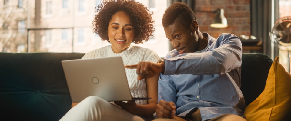 Man pointing at a laptop screen while talking to his girlfriend as they sit together on a sofa in a stylish loft apartment—capturing a shared moment during their apartment search.