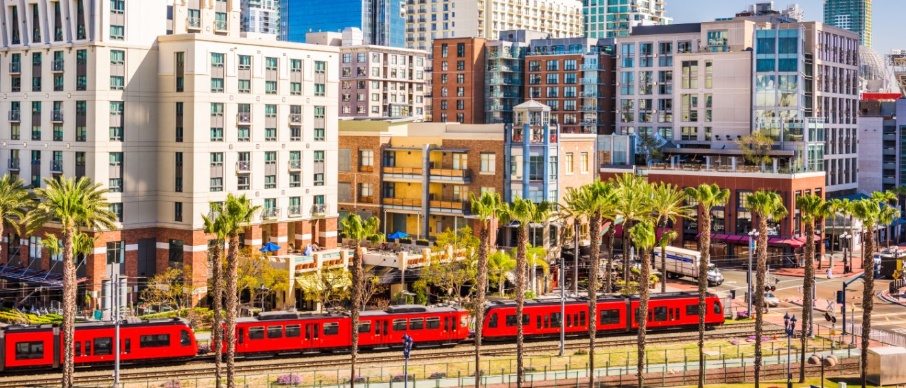 San Diego Trolley train passing through downtown, surrounded by palm trees and modern apartment buildings under a sunny sky.